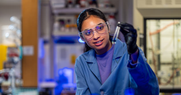 A girl in a lab coat, safety glasses, and gloves holding up a test tube. A lab is visible in the background