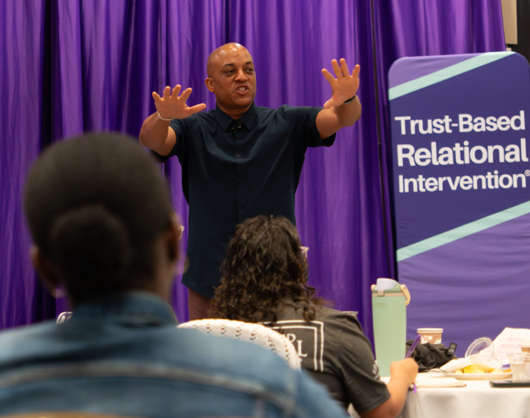   A speaker standing in front of a purple curtain addresses an audience with hands raised. A sign reads 